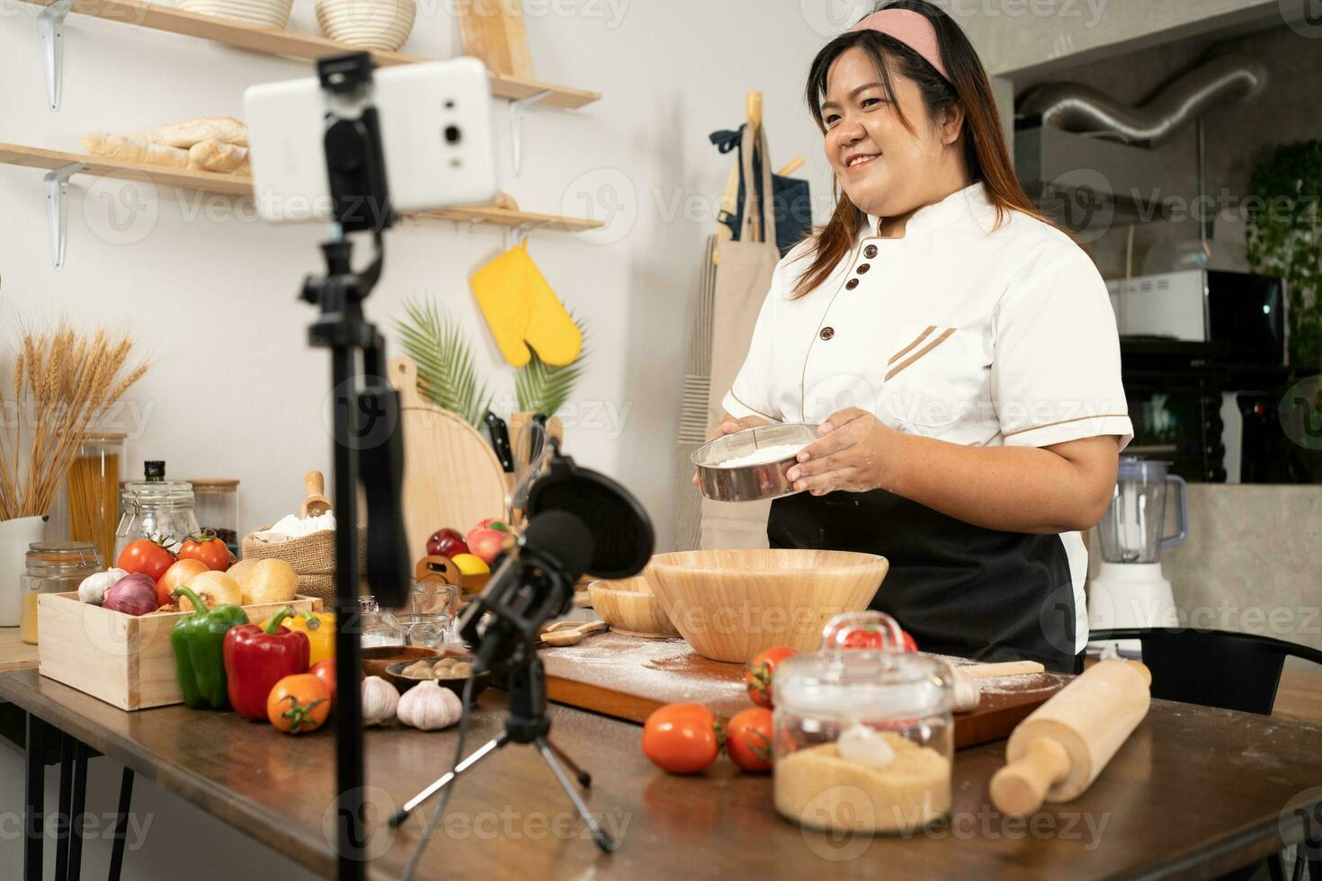 Young attractive Asian woman is blogging for her kitchen channel about homemade pizza while streaming online for webinar masterclass lesson. Vlogger broadcasting live video online teaching cooking photo