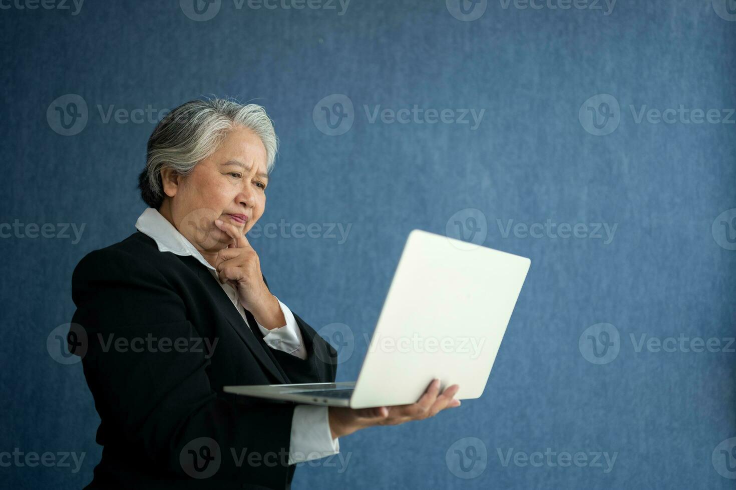 Portrait of intelligent senior businesswoman CEO manager holding in hands laptop creating a presentation on isolated blue background. Old Woman thinking after getting financial statements reports. photo