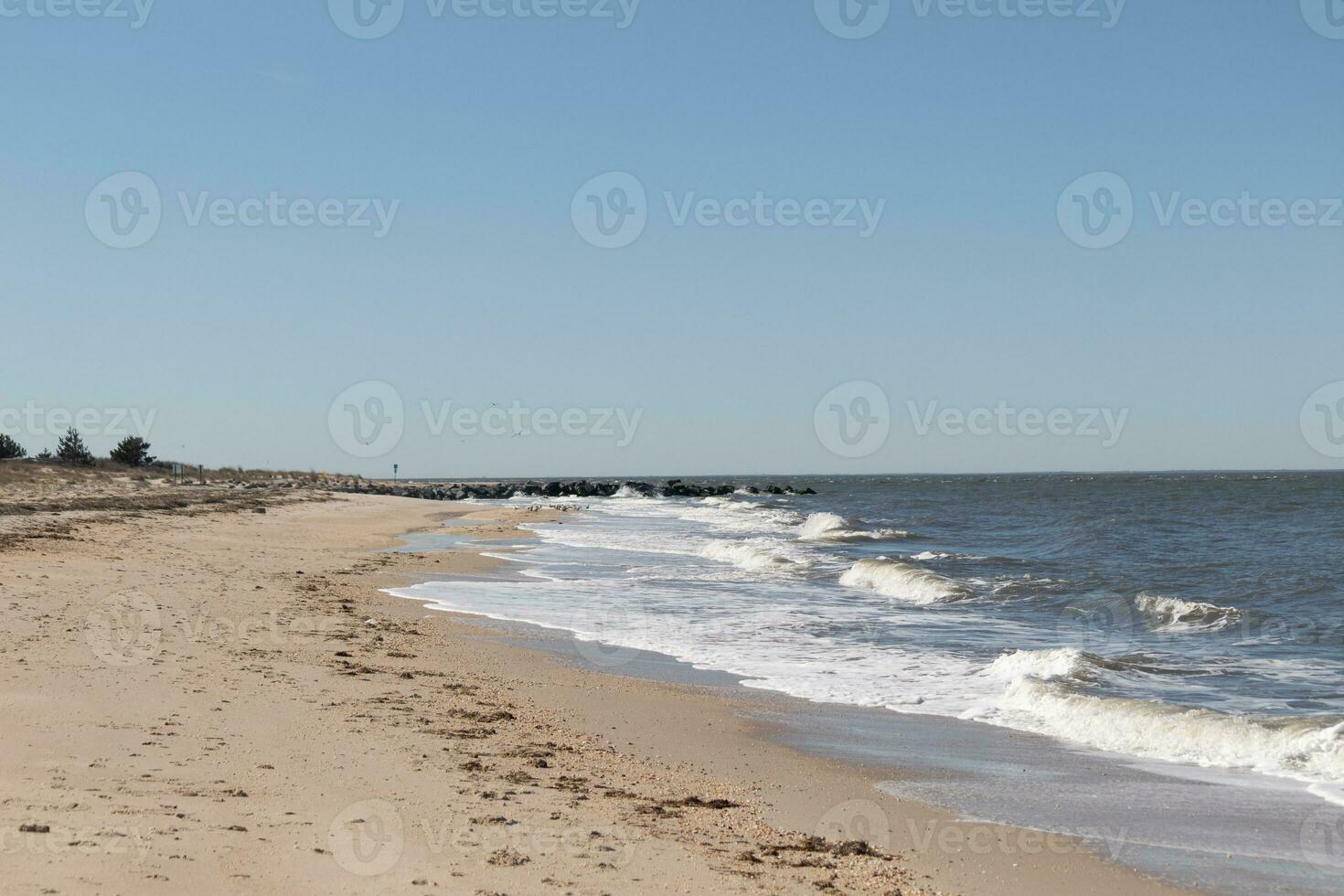 This beautiful image of the surf coming into the beach. The blue ocean with whitecaps on the waves. The water is pummeling the pretty brown sand. The sky is clear with hardly any clouds. photo