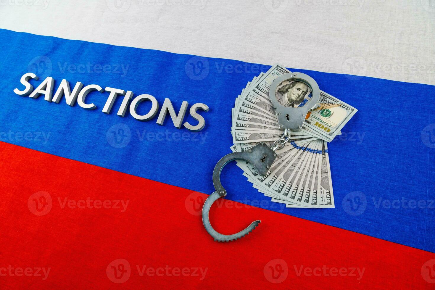the word sanctions laid with silver metal letters on russian tricolor flag near dollar banknotes and handcuffs in diagonal perspective photo