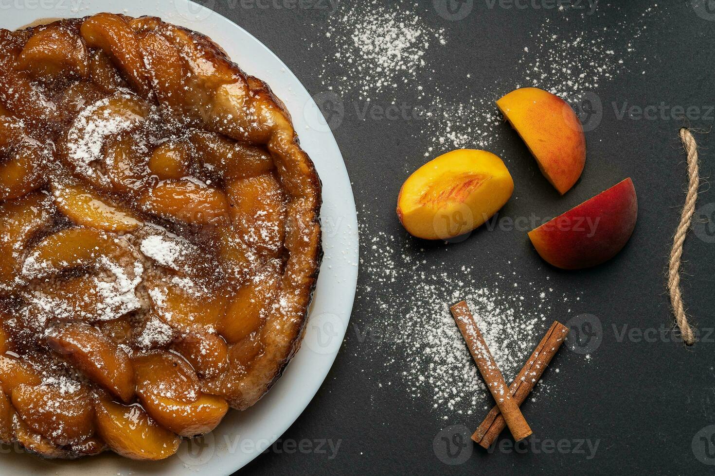 Tarte Tatin with peaches, caramel and powdered sugar on a white plate on a dark background. Near slices of peach and cinnamon sticks. Top view, horizontal orientation photo