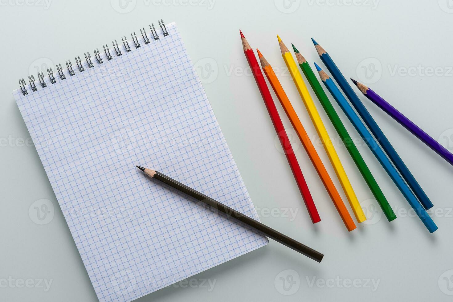 Clean notepad in a cage with a pencil lying on it and rainbow color pencils on a white background top view photo