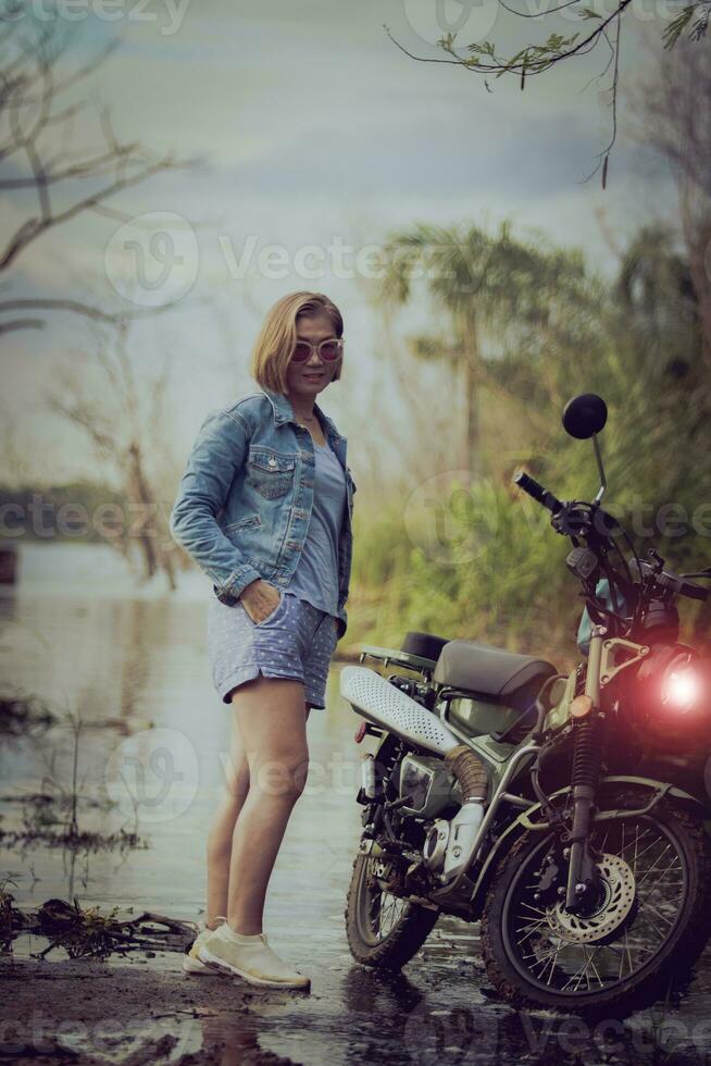 beautiful woman wearing blue jeans jacket standing beside enduro motorcycle against colorful natural  background photo