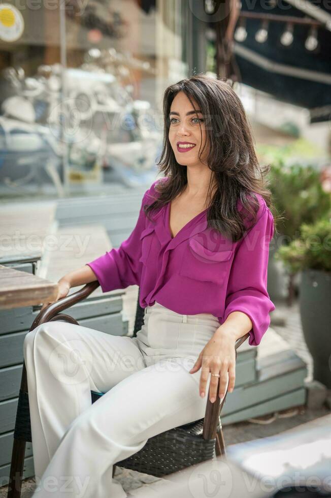 Happy Brunette Girl with pink blouse Sitting at the bar , Drinking a glass of lemonade While Smiling at the camera. Young sexy pretty woman on the chair  drinking  juice wearing a white pants photo