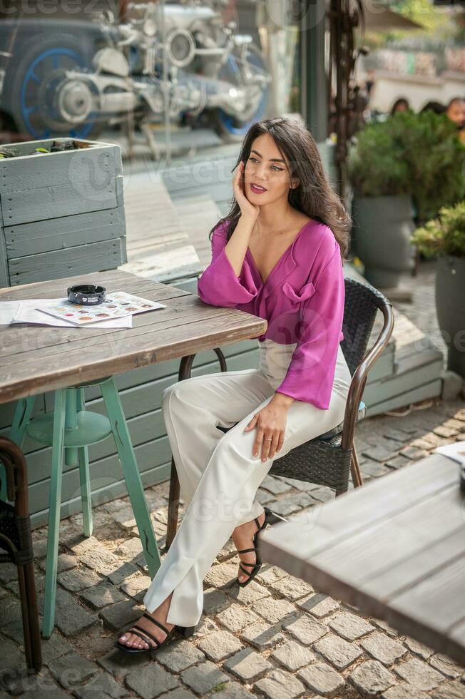 Happy Brunette Girl with pink blouse Sitting at the bar , Drinking a glass of lemonade While Smiling at the camera. Young sexy pretty woman on the chair  drinking  juice wearing a white pants photo