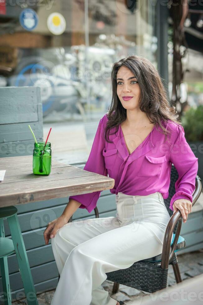 contento morena niña con rosado blusa sentado a el bar , Bebiendo un vaso de limonada mientras sonriente a el cámara. joven sexy bonito mujer en el silla Bebiendo jugo vistiendo un blanco pantalones foto