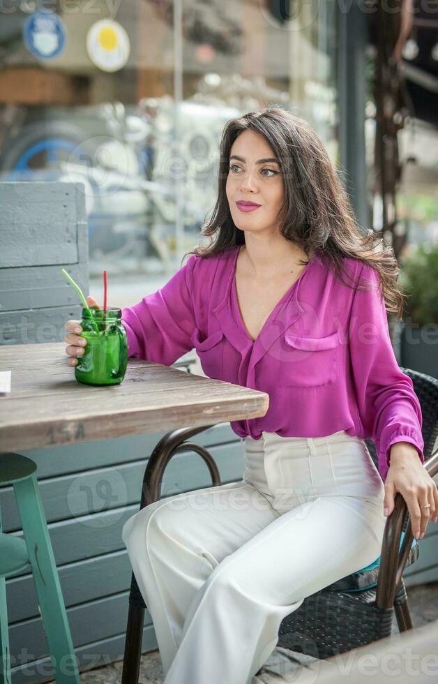 Happy Brunette Girl with pink blouse Sitting at the bar , Drinking a glass of lemonade While Smiling at the camera. Young sexy pretty woman on the chair  drinking  juice wearing a white pants photo