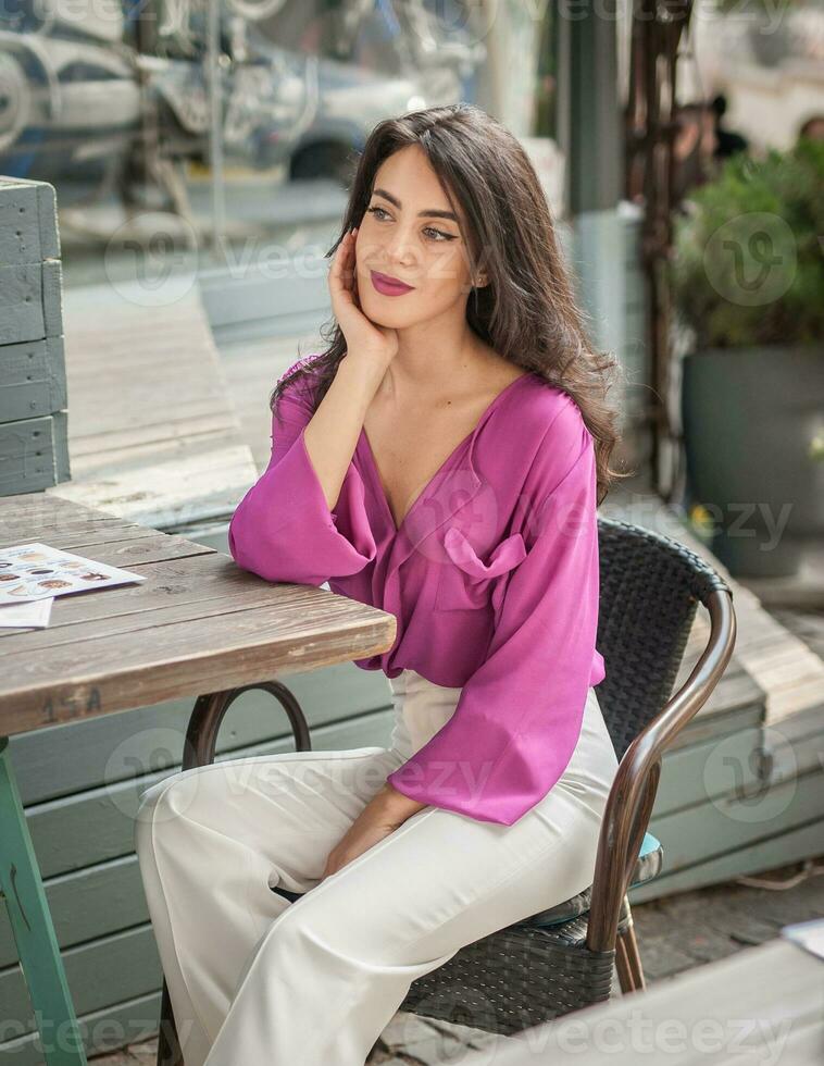Happy Brunette Girl with pink blouse Sitting at the bar , Drinking a glass of lemonade While Smiling at the camera. Young sexy pretty woman on the chair  drinking  juice wearing a white pants photo