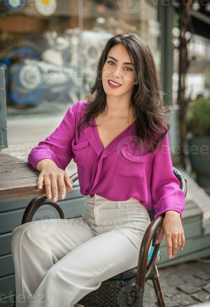 Happy Brunette Girl with pink blouse Sitting at the bar , Drinking a glass of lemonade While Smiling at the camera. Young sexy pretty woman on the chair  drinking  juice wearing a white pants photo