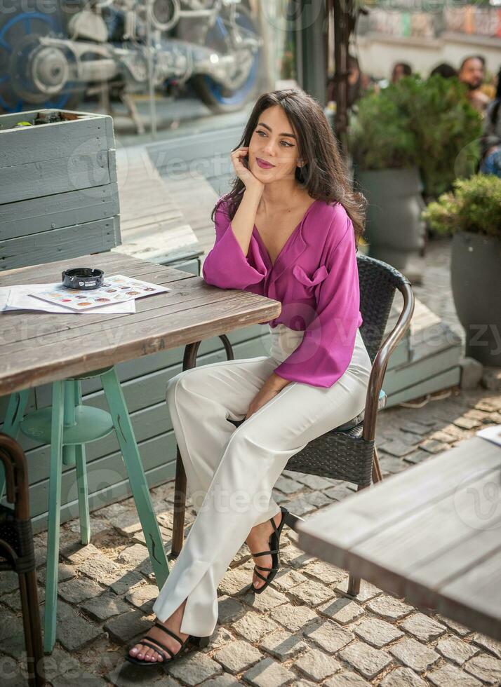 Happy Brunette Girl with pink blouse Sitting at the bar , Drinking a glass of lemonade While Smiling at the camera. Young sexy pretty woman on the chair  drinking  juice wearing a white pants photo