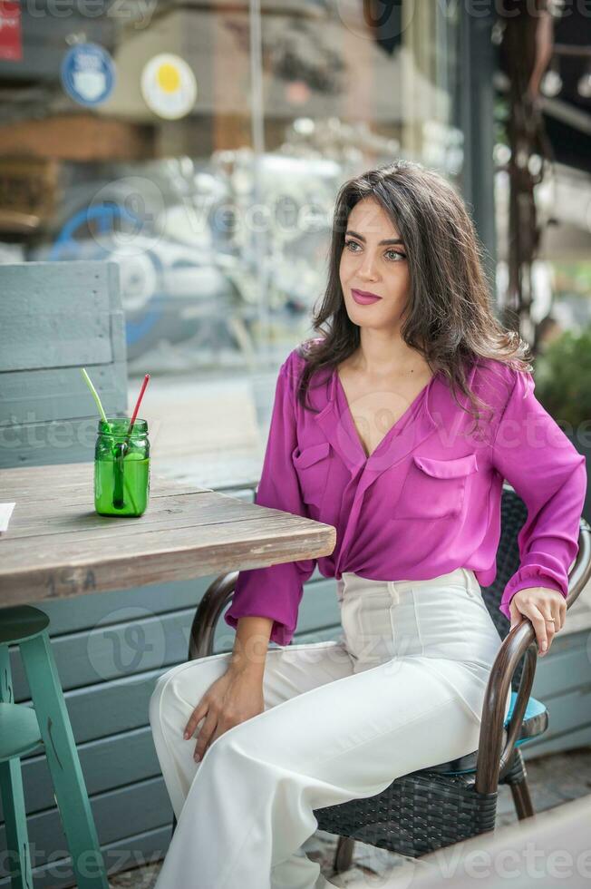Happy Brunette Girl with pink blouse Sitting at the bar , Drinking a glass of lemonade While Smiling at the camera. Young sexy pretty woman on the chair  drinking  juice wearing a white pants photo