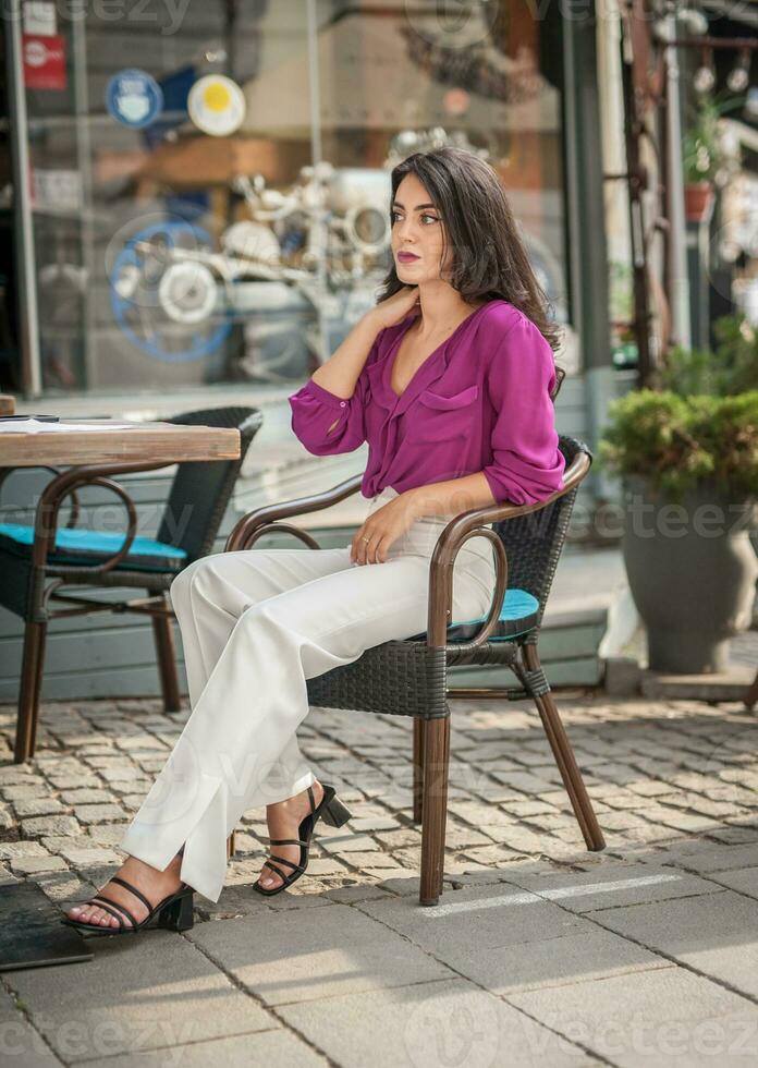 contento morena niña con rosado blusa sentado a el bar , Bebiendo un vaso de limonada mientras sonriente a el cámara. joven sexy bonito mujer en el silla Bebiendo jugo vistiendo un blanco pantalones foto