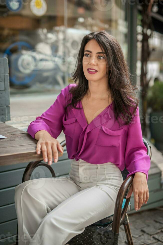 Happy Brunette Girl with pink blouse Sitting at the bar , Drinking a glass of lemonade While Smiling at the camera. Young sexy pretty woman on the chair  drinking  juice wearing a white pants photo