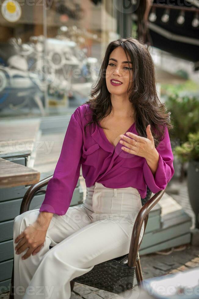 Happy Brunette Girl with pink blouse Sitting at the bar , Drinking a glass of lemonade While Smiling at the camera. Young sexy pretty woman on the chair  drinking  juice wearing a white pants photo
