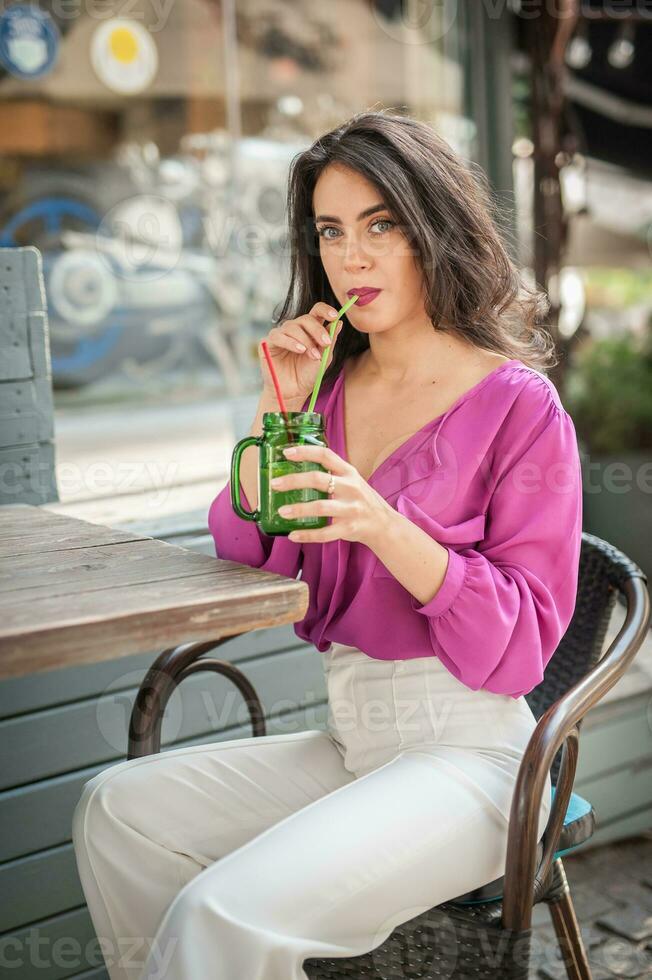 contento morena niña con rosado blusa sentado a el bar , Bebiendo un vaso de limonada mientras sonriente a el cámara. joven sexy bonito mujer en el silla Bebiendo jugo vistiendo un blanco pantalones foto