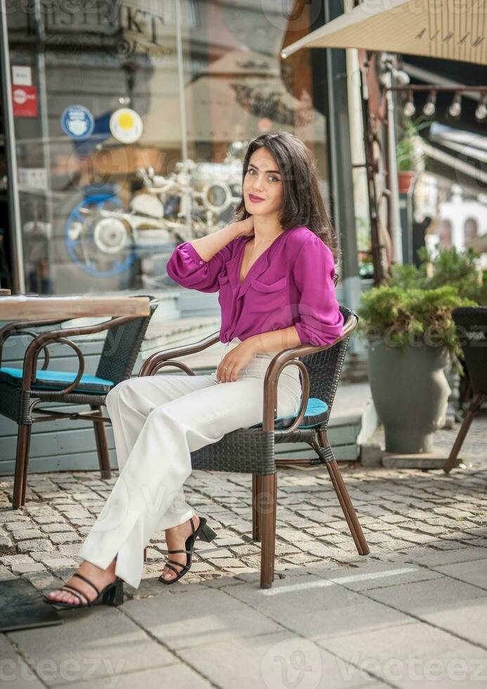 contento morena niña con rosado blusa sentado a el bar , Bebiendo un vaso de limonada mientras sonriente a el cámara. joven sexy bonito mujer en el silla Bebiendo jugo vistiendo un blanco pantalones foto