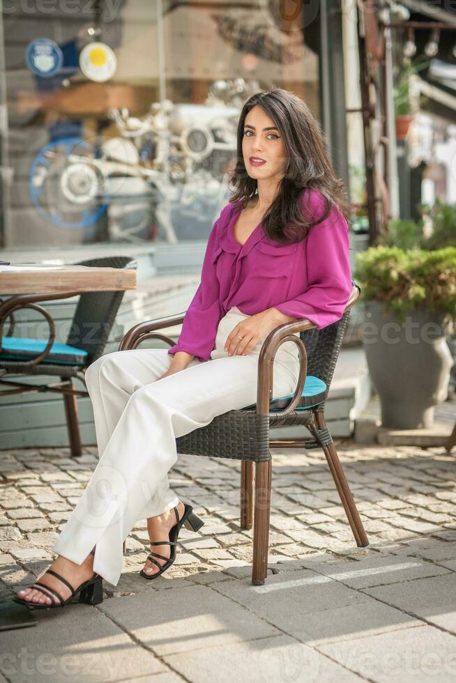 contento morena niña con rosado blusa sentado a el bar , Bebiendo un vaso de limonada mientras sonriente a el cámara. joven sexy bonito mujer en el silla Bebiendo jugo vistiendo un blanco pantalones foto