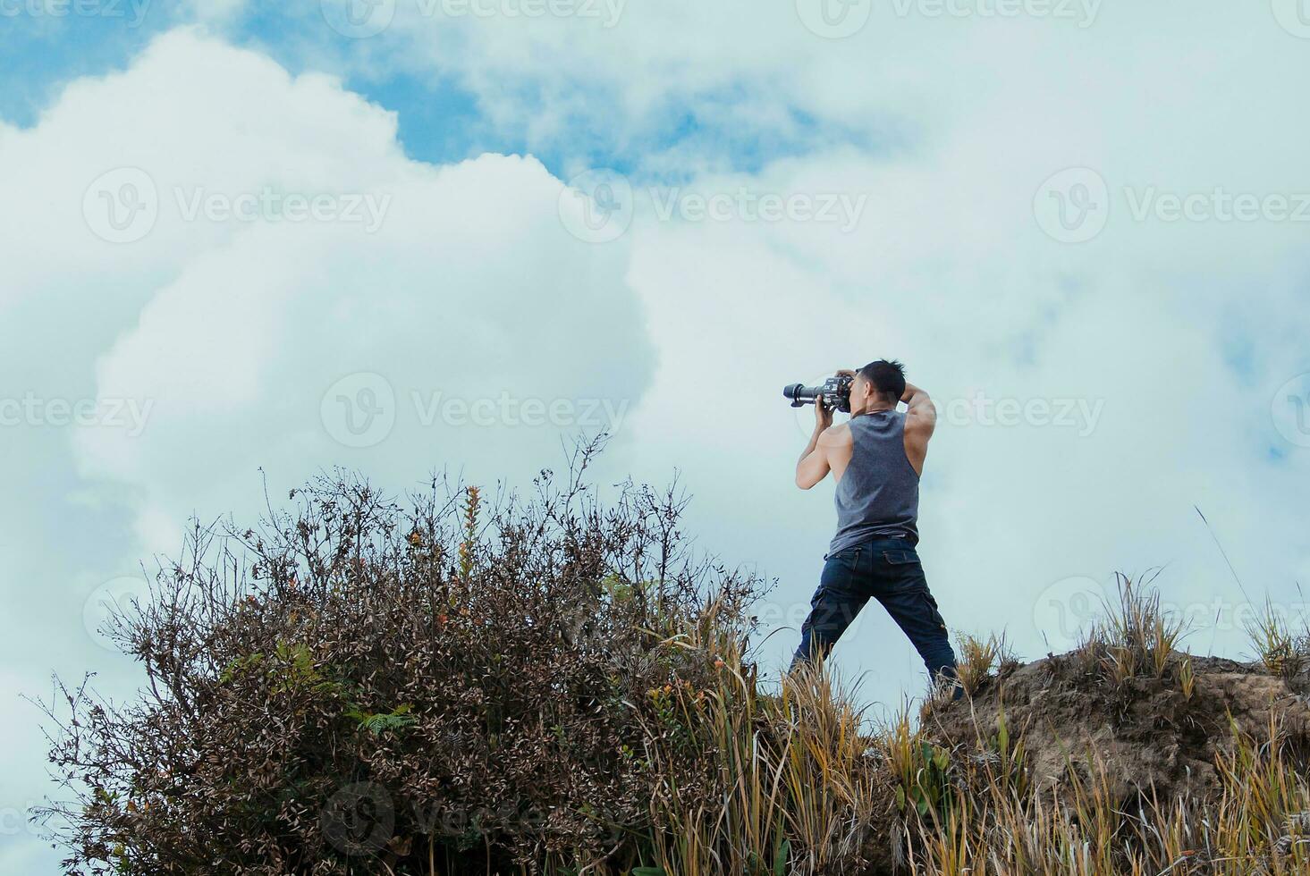 Indonesian male photographer wearing a singlet on a hill with clouds and sky in the background. photo