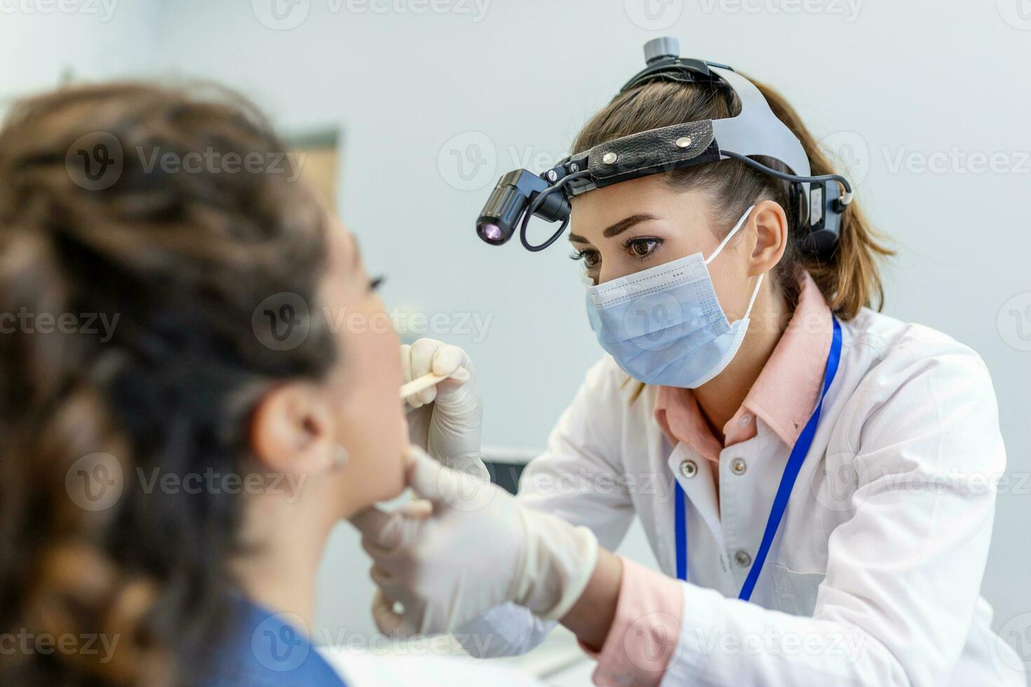 A young woman sits on an exam table across from her doctor. The doctor reaches forward with a tongue depressor as the woman looks up and sticks out her tongue. photo