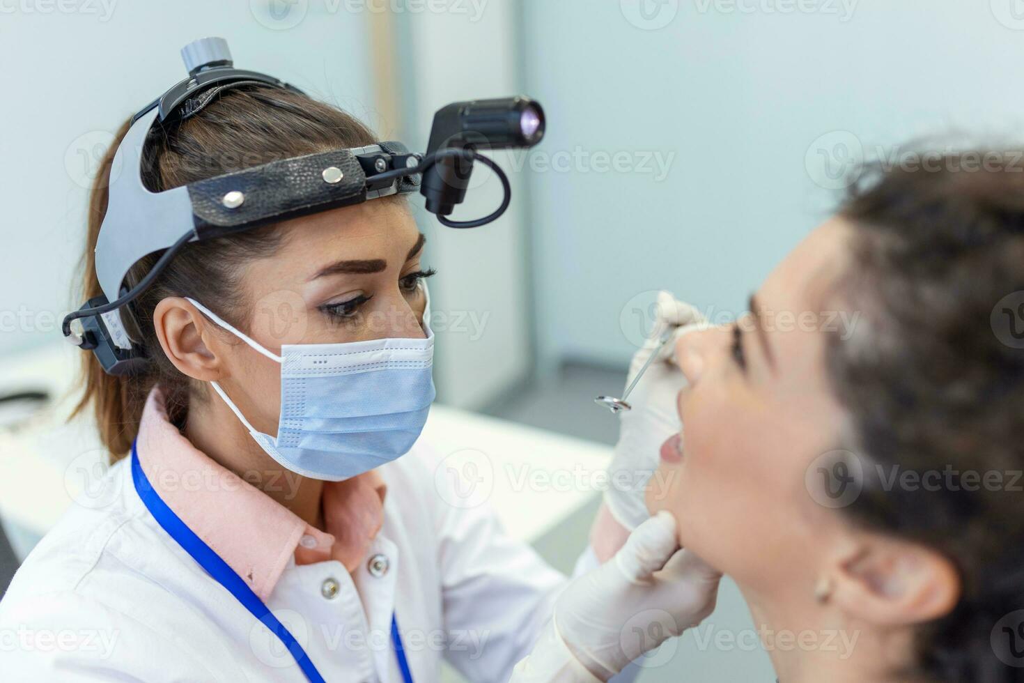 Female patient opening her mouth for the doctor to look in her throat. Female doctor examining sore throat of patient in clinic. Otolaryngologist examines sore throat of patient. photo