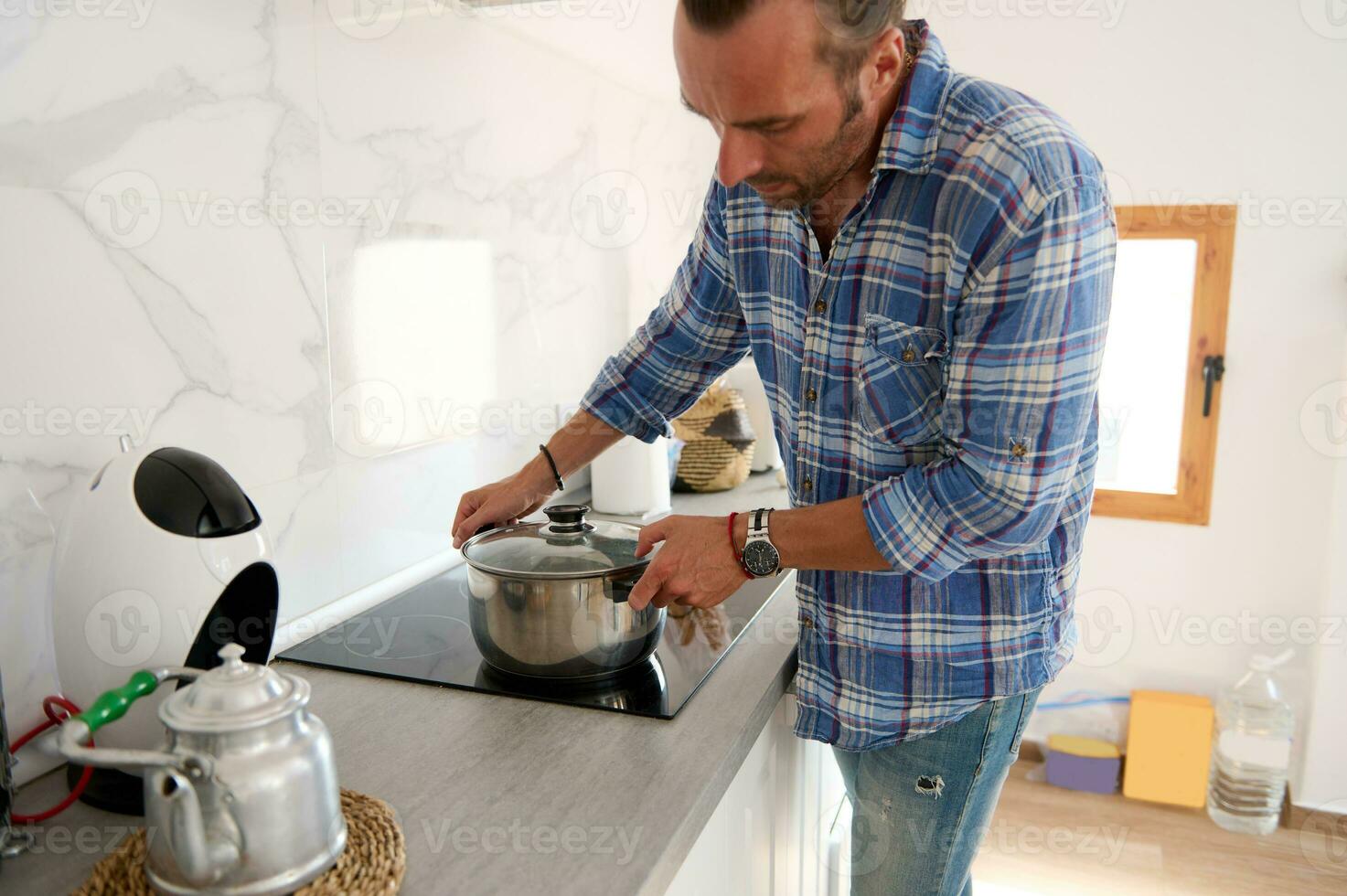 Young adult Caucasian handsome young man putting a saucepan on an electric stove while cooking dinner in home kitchen photo