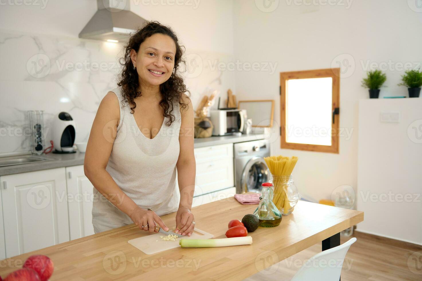 Beautiful woman housewife smiles looking at camera, chopping leek and garlic on cutting board in minimalist home kitchen photo