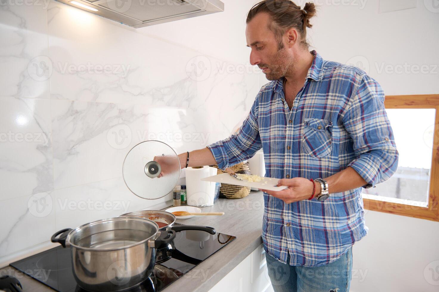Male chef putting chopped garlic from cutting board into a saucepan with boiling tomato sauce, preparing dinner photo