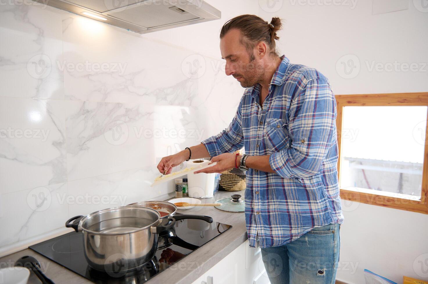 Handsome young man putting chopped garlic from cutting board into a saucepan with boiling tomato sauce, cooking at home photo