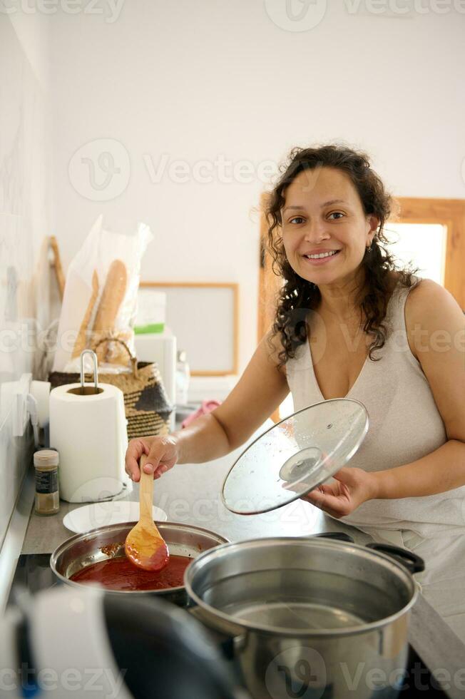 auténtico bonito mujer, un ama de casa sonriente mirando a cámara, preparando un delicioso cena en un minimalista cocina foto