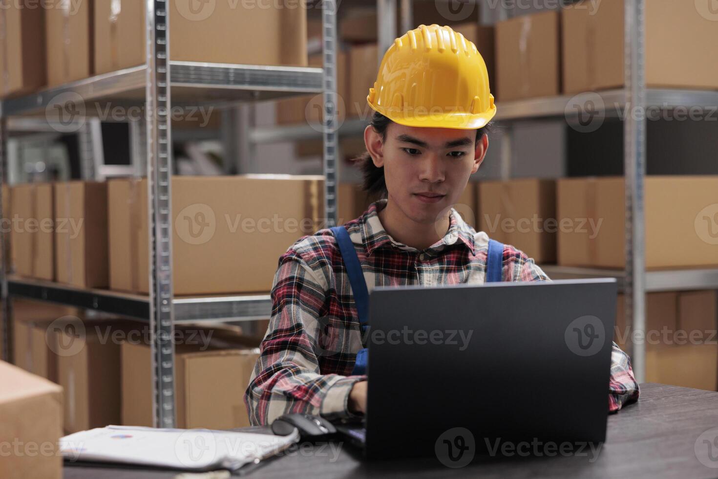 Young asian man warehouse operator managing delivery on laptop while working at storage room desk. Storehouse manager in yellow helmet analyzing products logistics on computer photo