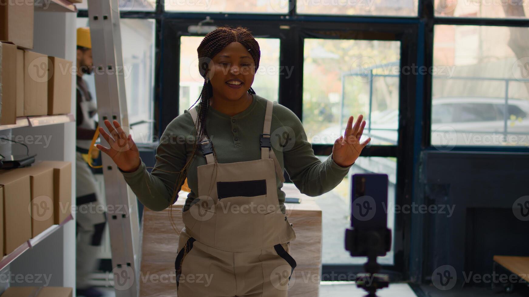 Warehouse chief supply chain officer using smartphone to make training video for interns. African american supervisor films herself in depository showing trainees how to seal cardboard boxes photo