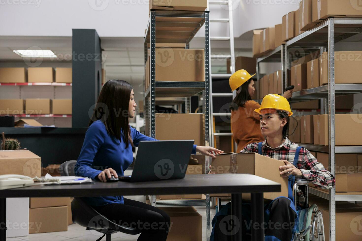 Warehouse manager coordinating logistics operators team from desk and checking parcel packing quality. Asian storehouse worker in wheelchair giving cardboard box to coworker at table photo