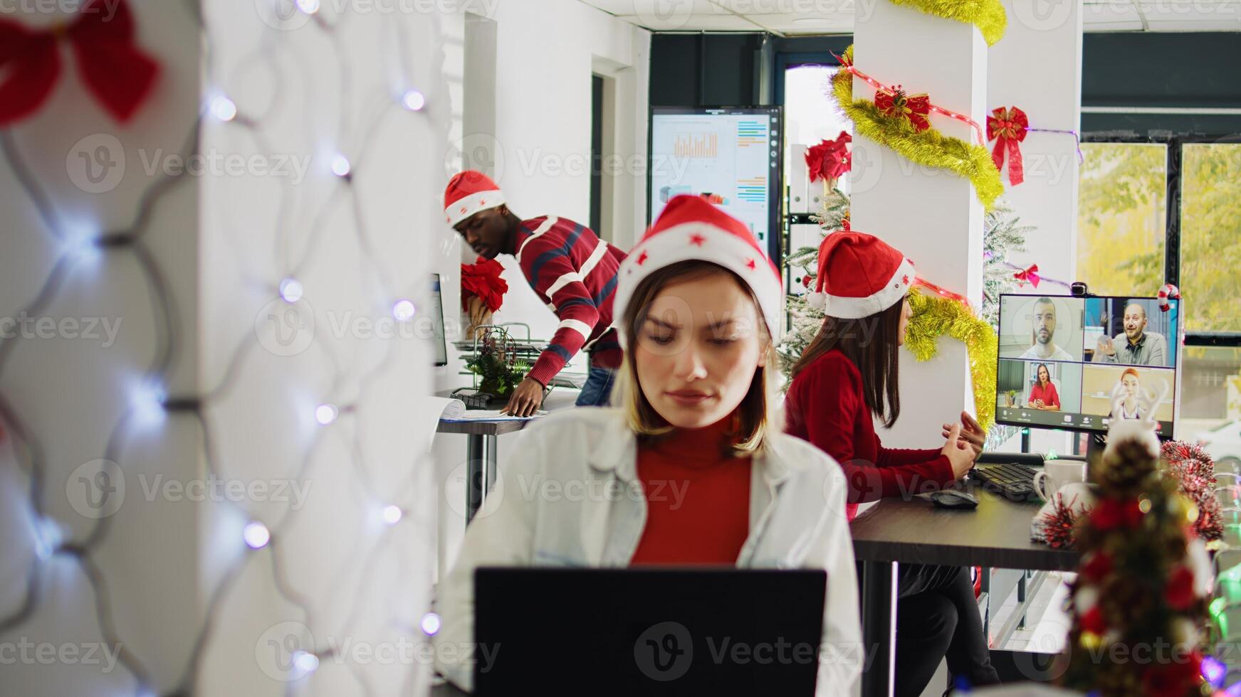 Asian staff member in videocall with teammates during Christmas season in festive decorated office. Employee in teleconference meeting with team in busy xmas ornate workplace photo