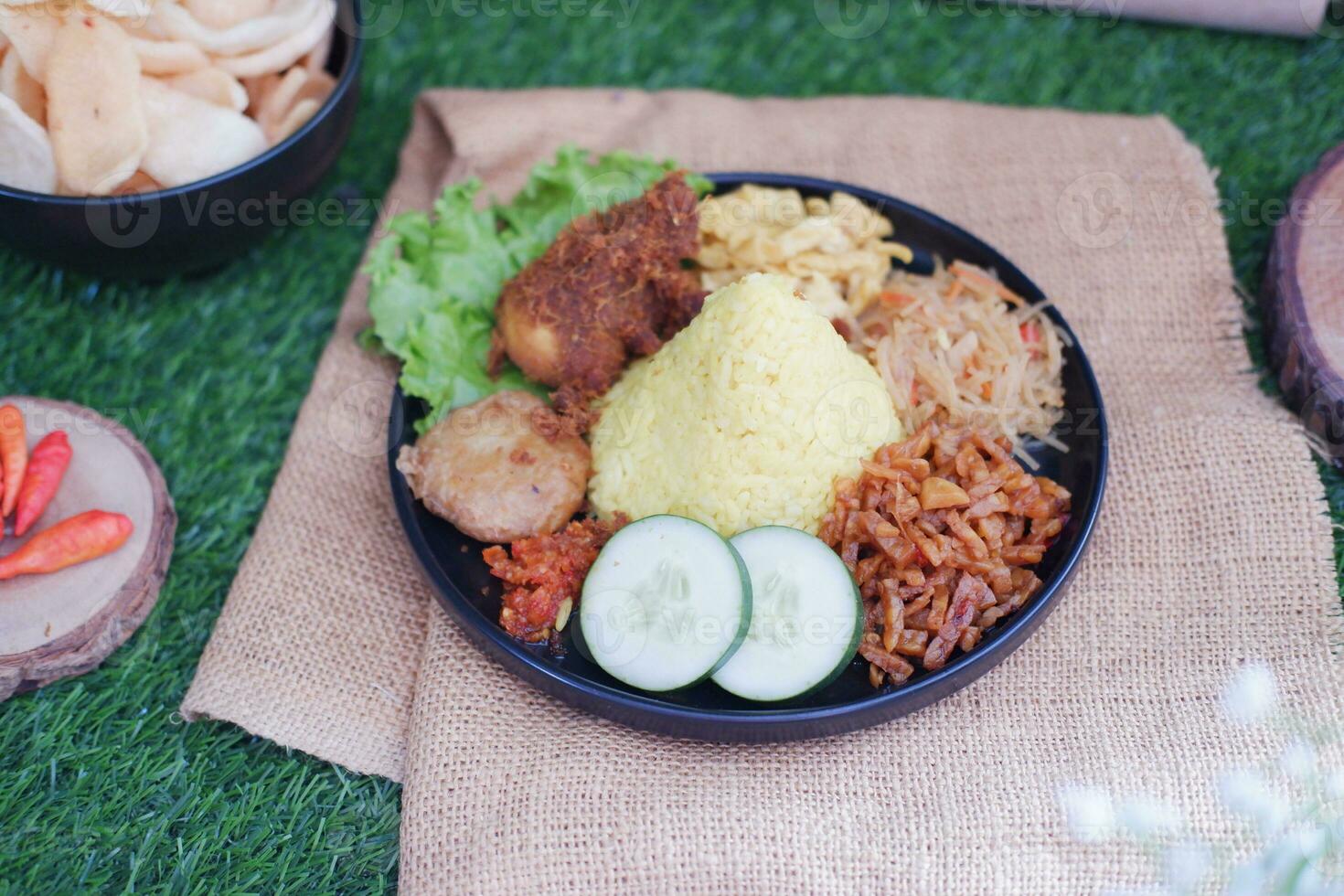 indonesian style yellow rice with chicken and side dishes in black plate on green grass background photo