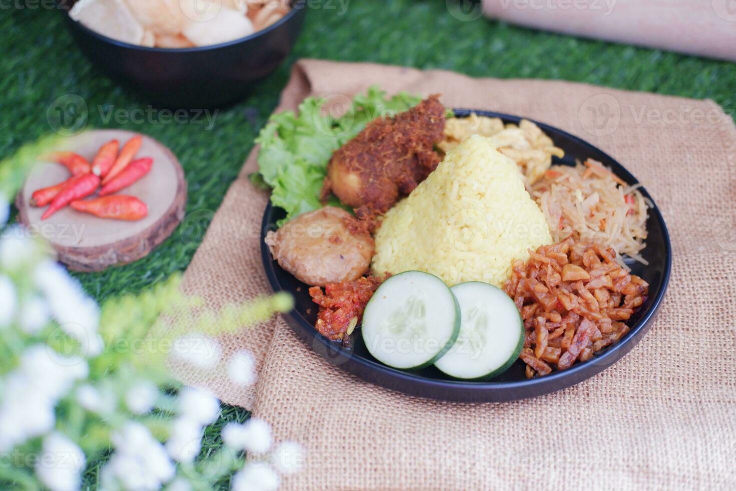 indonesian style yellow rice with chicken and side dishes in black plate on green grass background photo