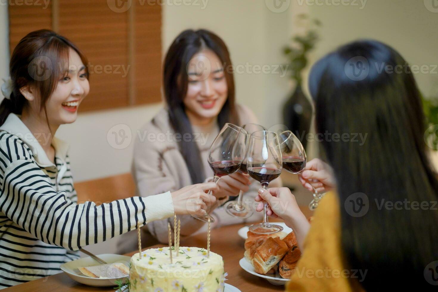 Cheerful friends enjoying home Birthday holiday party. Asian sister cheering drinking red wine celebrating with Birthday cake. photo