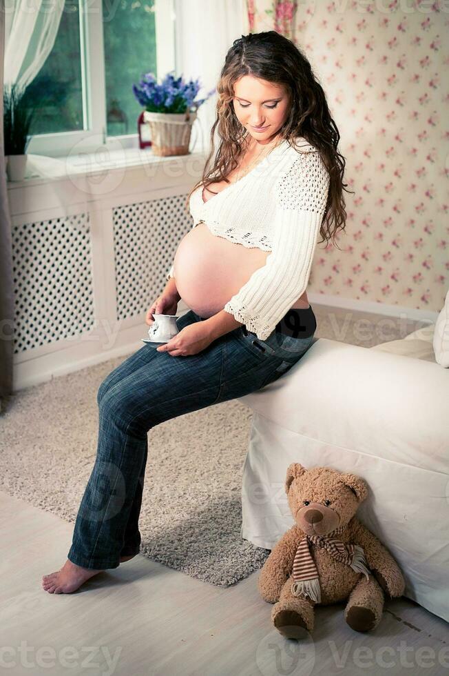 A pregnant woman in jeans and a skimpy jacket sitting on a sofa next to a teddy bear photo