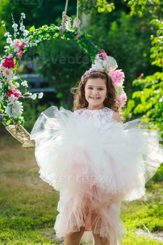 hermosa niña en un elegante rosado vestir saltos cerca columpio decorado con flores niño celebra cumpleaños 8 años. hermosa flores primavera tema columpio en jardín foto