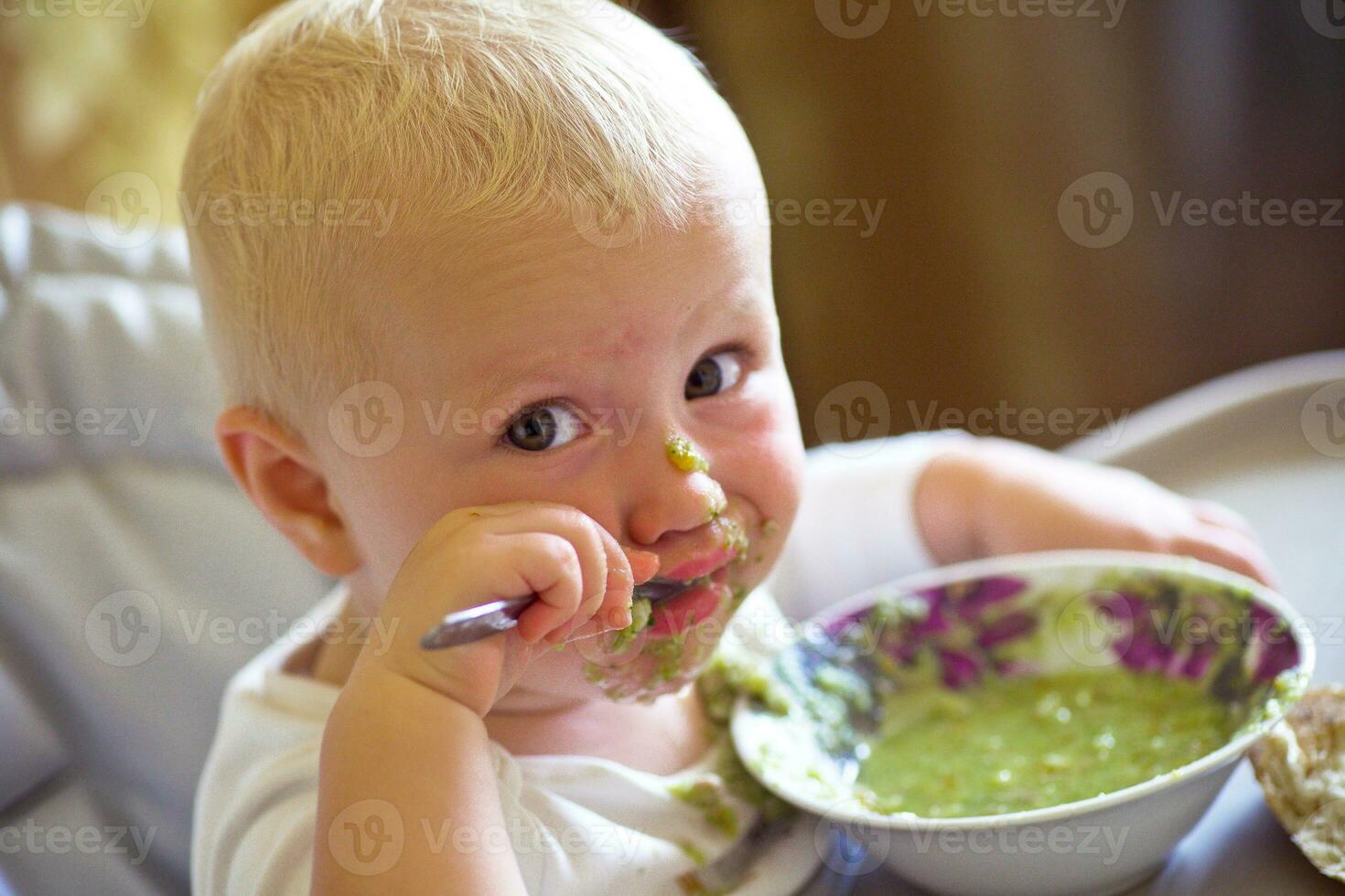 A little girl is eating broccoli soup while sitting in a high chair in the kitchen photo