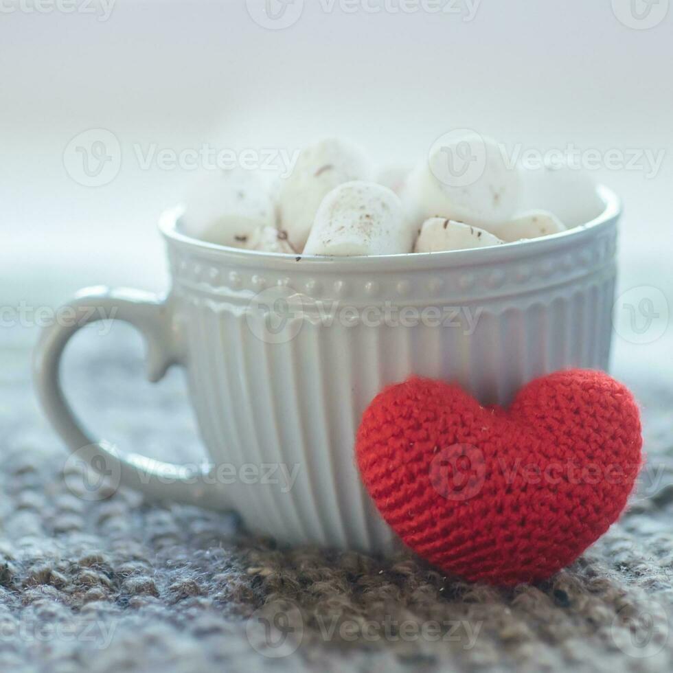 A big crochet red heart next to a cup of cocoa. Cappuccino in a gray cup against a white background. photo