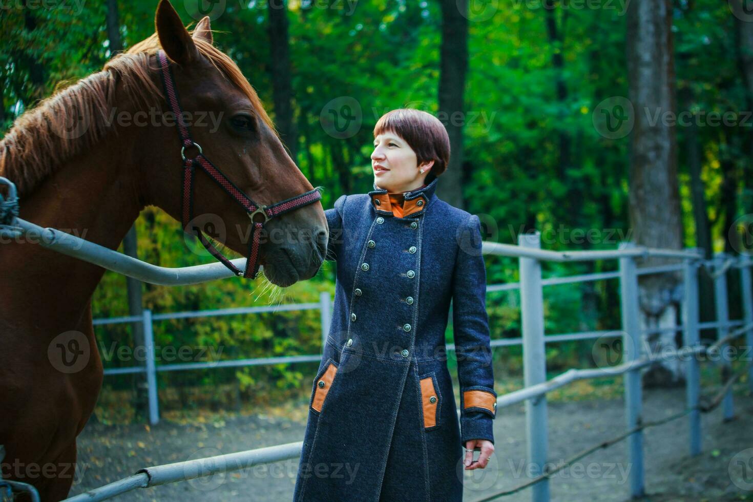 A woman in a long blue coat strokes the horse's face against the background of a green forest. A woman stands at an iron fence enclosing a pen for horses. photo