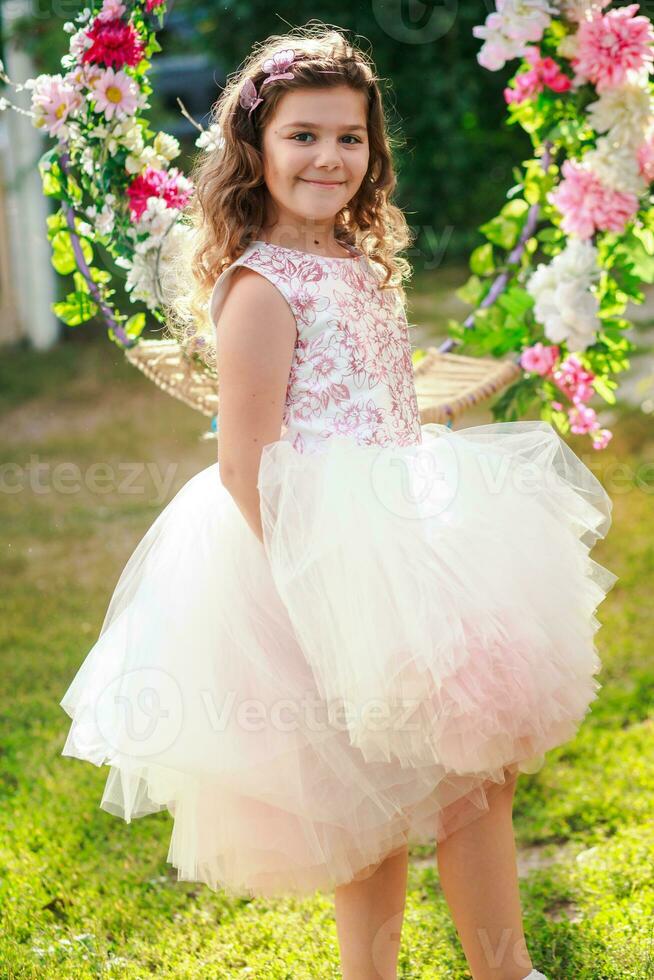 hermosa niña en un elegante rosado vestir saltos cerca columpio decorado con flores niño celebra cumpleaños 8 años. hermosa flores primavera tema columpio en jardín foto