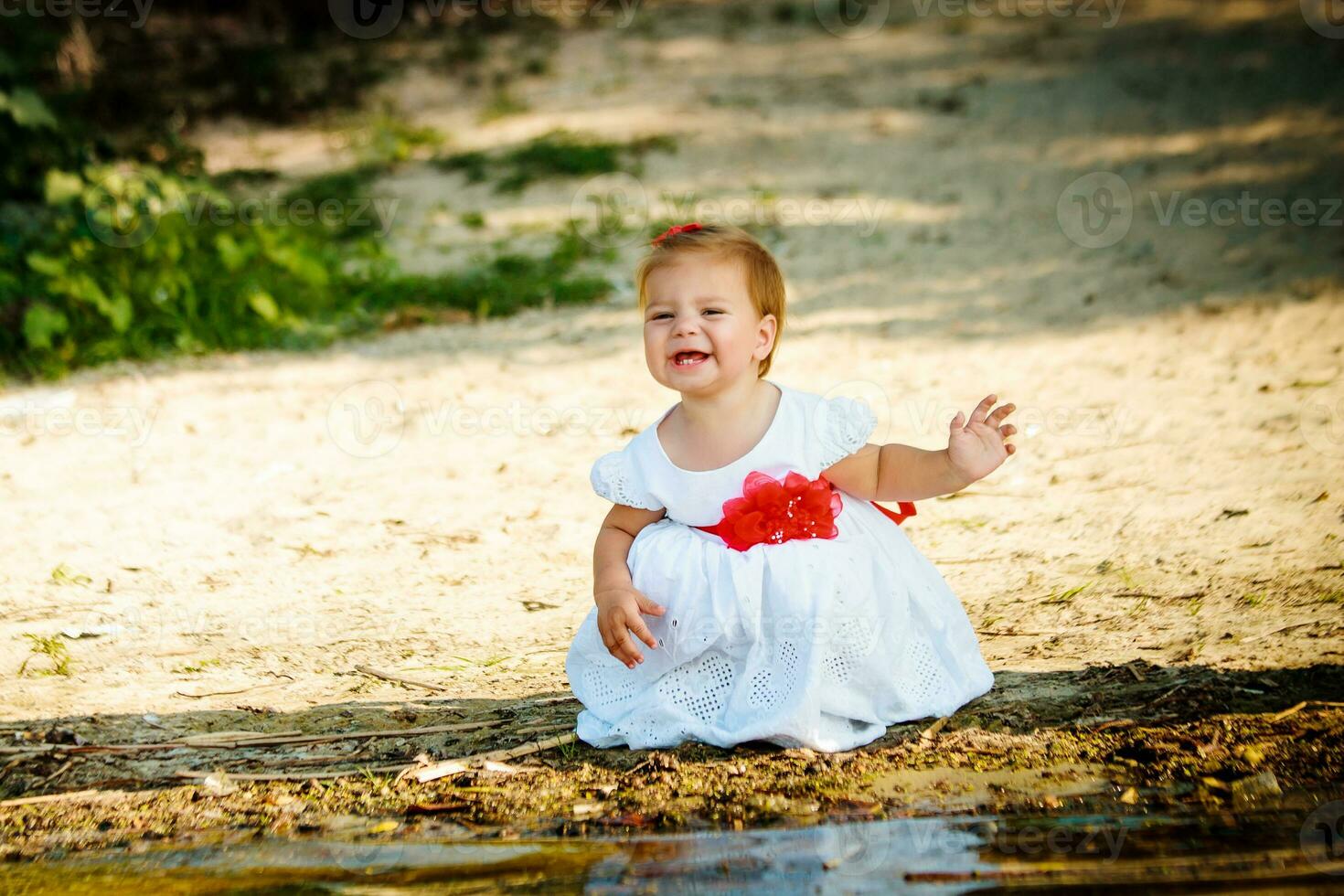 The little girl in a white dress sitting on the river bank. A child playing near the water. The kid smiles. Child grimaced and closed his eyes. photo