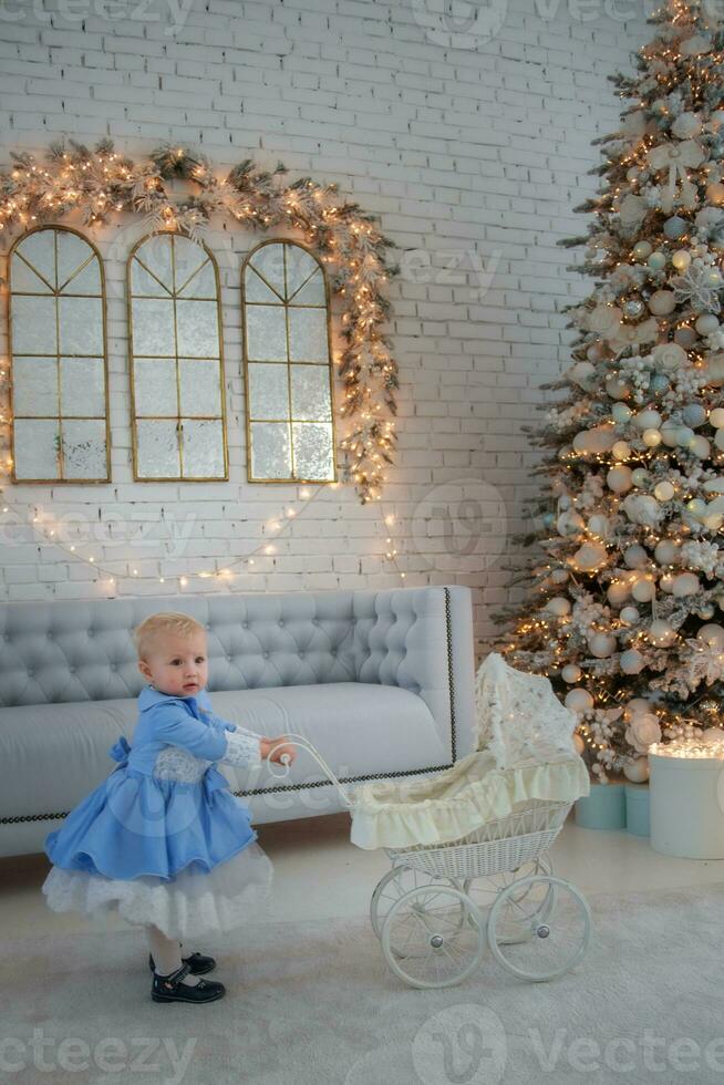 Baby girl wearing cute dress and headband, carries stroller in festively decorated room with garland of lights. photo