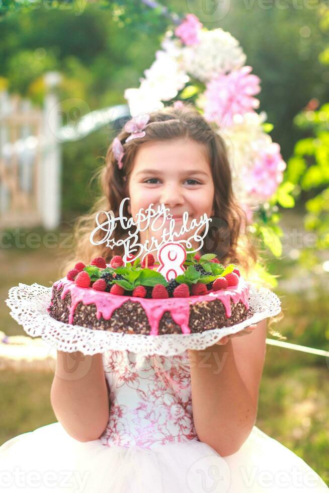 girl holding birthday cake with number 8 and inscription Happy Birthday. Celebrating ceremonial events on street during quarantine. child in background decorated photo zone of flowers.