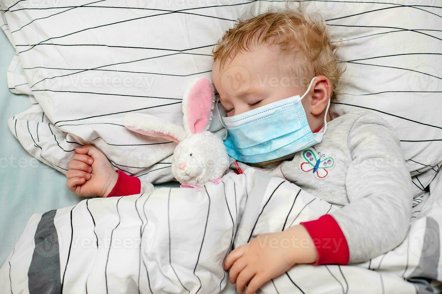 little child sleeps in bed in protective medical mask. sick child in white bed. Kid toddler one years old with flu, influenza or cold protected from viruses, pollution among patients with coronavirus photo