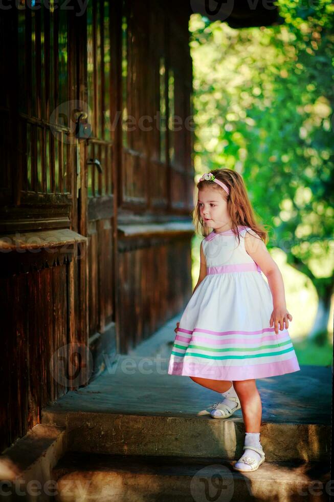 el niña abajo el escalera de un de madera casa en el aldea. hermosa de madera puerta en un antiguo casa. un niño en un hermosa blanco vestir caminando en el calle. foto