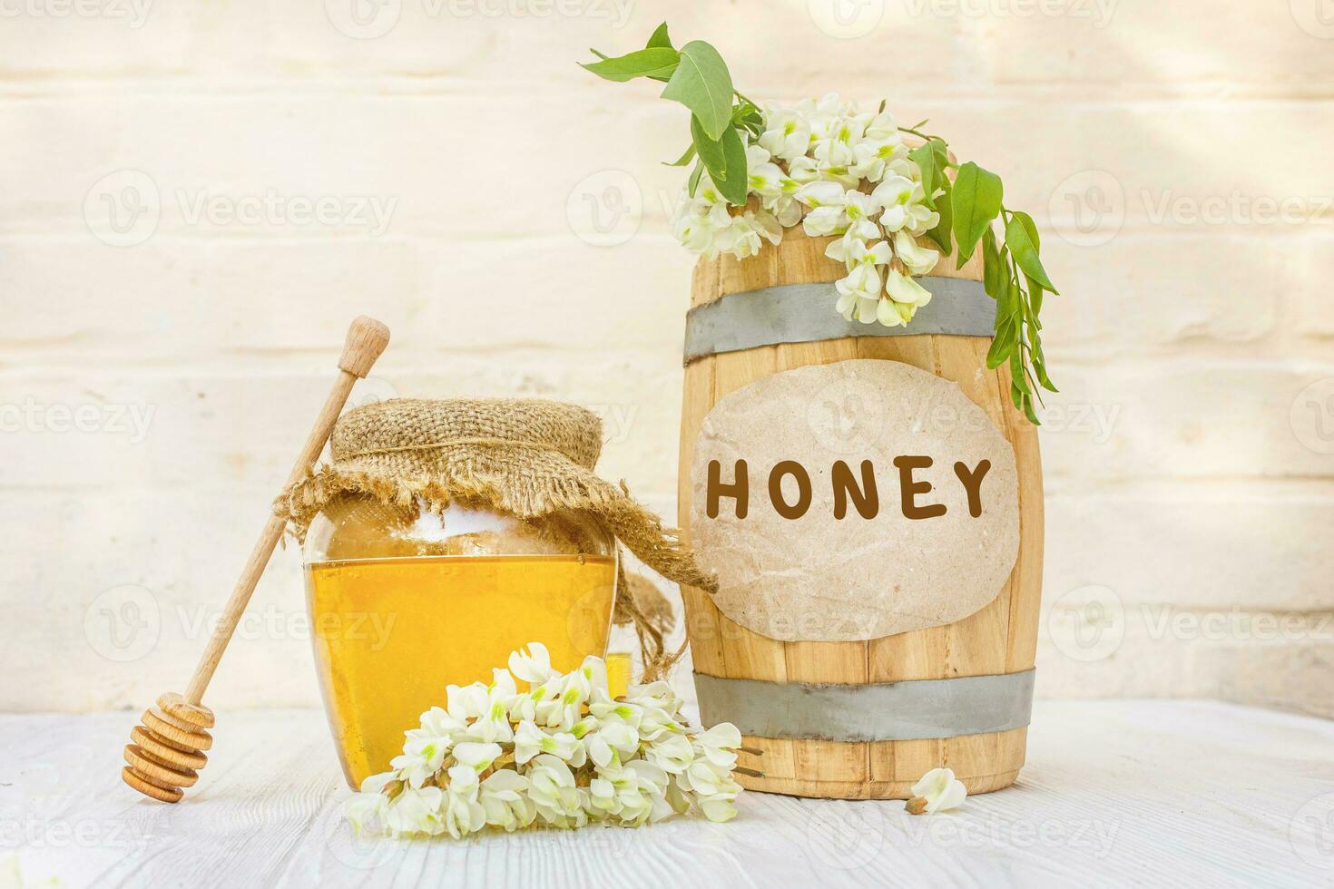 Text HONEY. Fresh liquid acacia honey harvest 2021 in a wooden barrel and a transparent jar with a canvas napkin on the lid. Branches of flowering white acacia photo