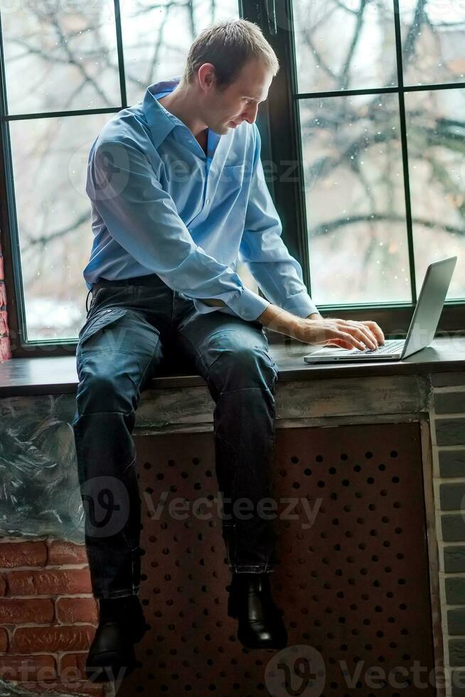 A man looks at the laptop screen and writes the message. The man is sitting on a windowsill near the window. Freelancer works at home in a comfortable environment. Soft focus photo