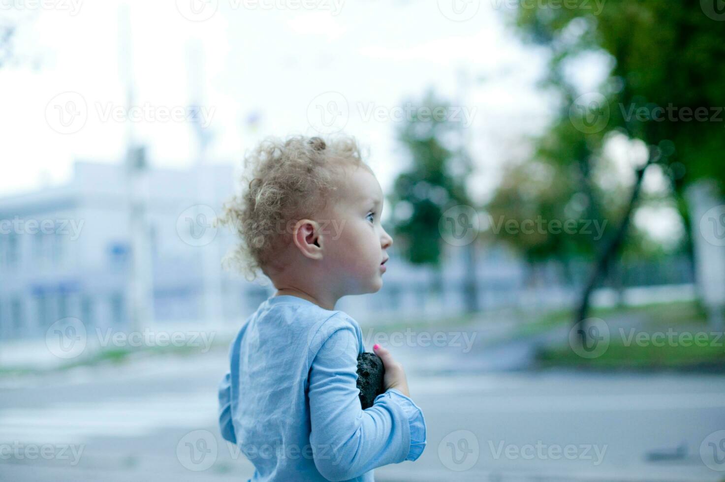 Little beautiful girl with short curly hair, walks around the city. The child holds a toy and is near the road. photo